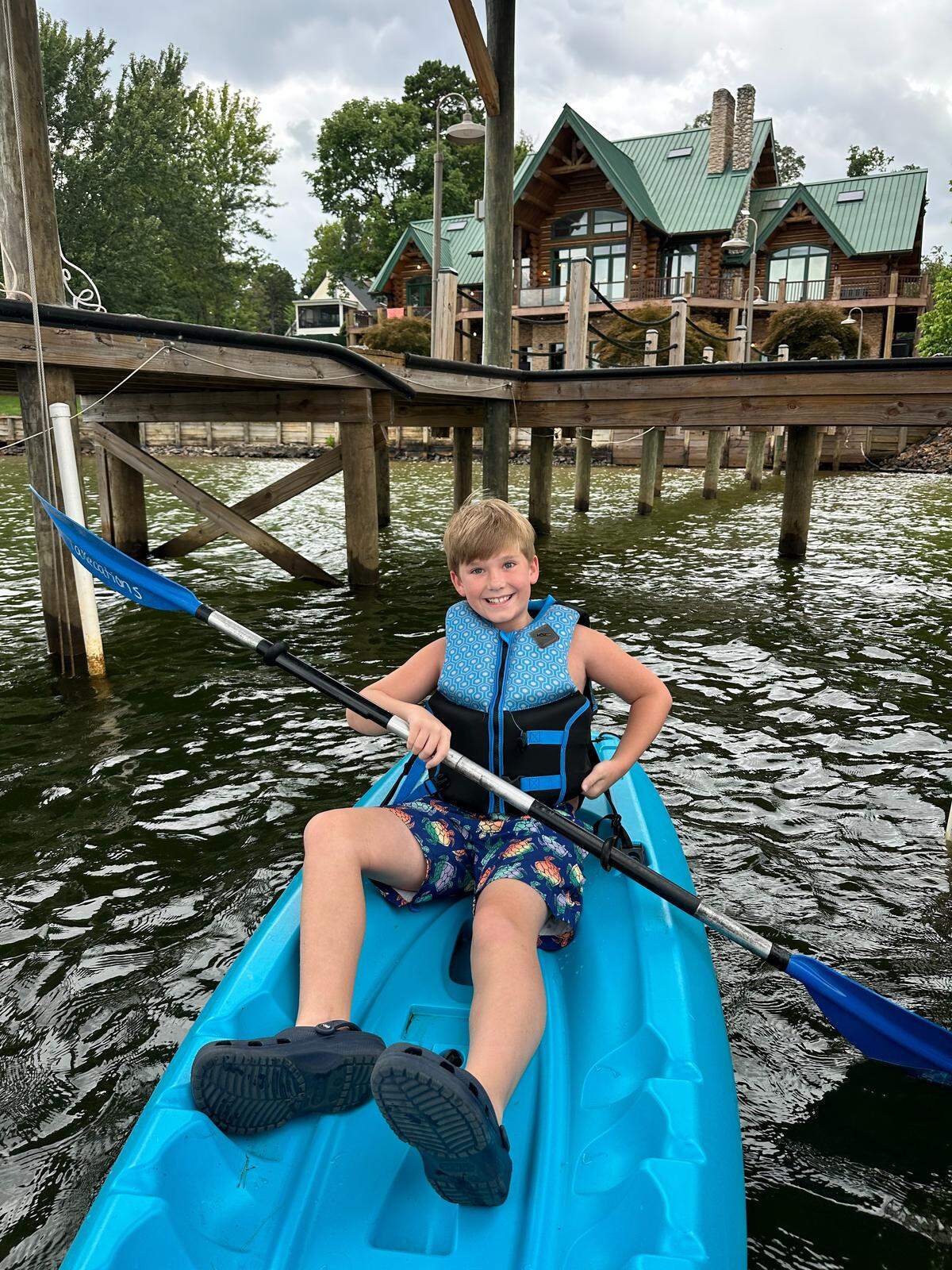 A child wearing a life vest paddles a bright blue kayak on a calm lake. In the background, a large, multi-story log cabin with a green roof sits on the shore.