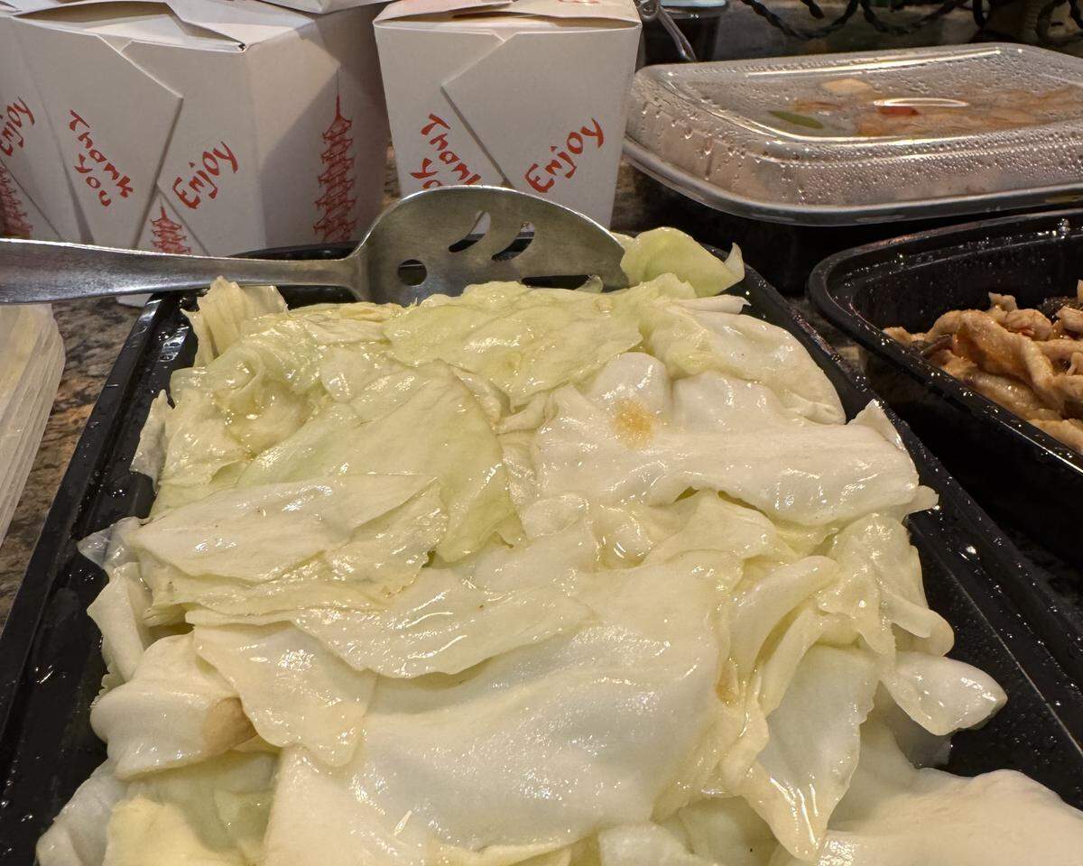 A dish of Taiwan sautéed cabbage from Lee Cafe is shown in a black plastic container. The tender, cooked cabbage is ready to be served with a large slotted spoon. Other takeout containers are visible in the background, indicating a to-go meal.