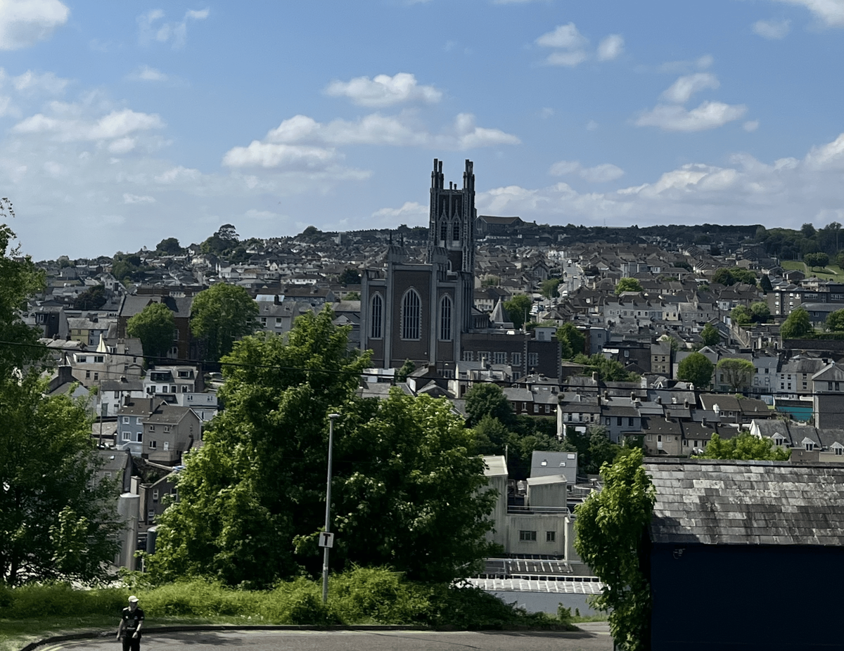 A view of a dense hillside city with a large, ornate church building with two tall steeples as the centerpiece. The city is made up of a mix of rooftops and buildings, and the entire scene is surrounded by green trees. In the foreground, a single person in a uniform is walking down a street. The sky is a bright blue with scattered white clouds.