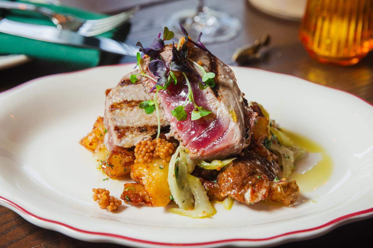 A close-up photograph of a seared tuna steak, sliced to show its pink-red rare center, stacked on a bed of golden-brown roasted potatoes and green vegetable slivers (possibly fennel or cabbage). The dish is garnished with whole-grain mustard seeds and purple microgreens. It is served on a white plate with a subtle wavy rim and a thin red edge. The background is softly blurred but shows a dark wooden table, a clear wine glass stem, and an orange textured glass.