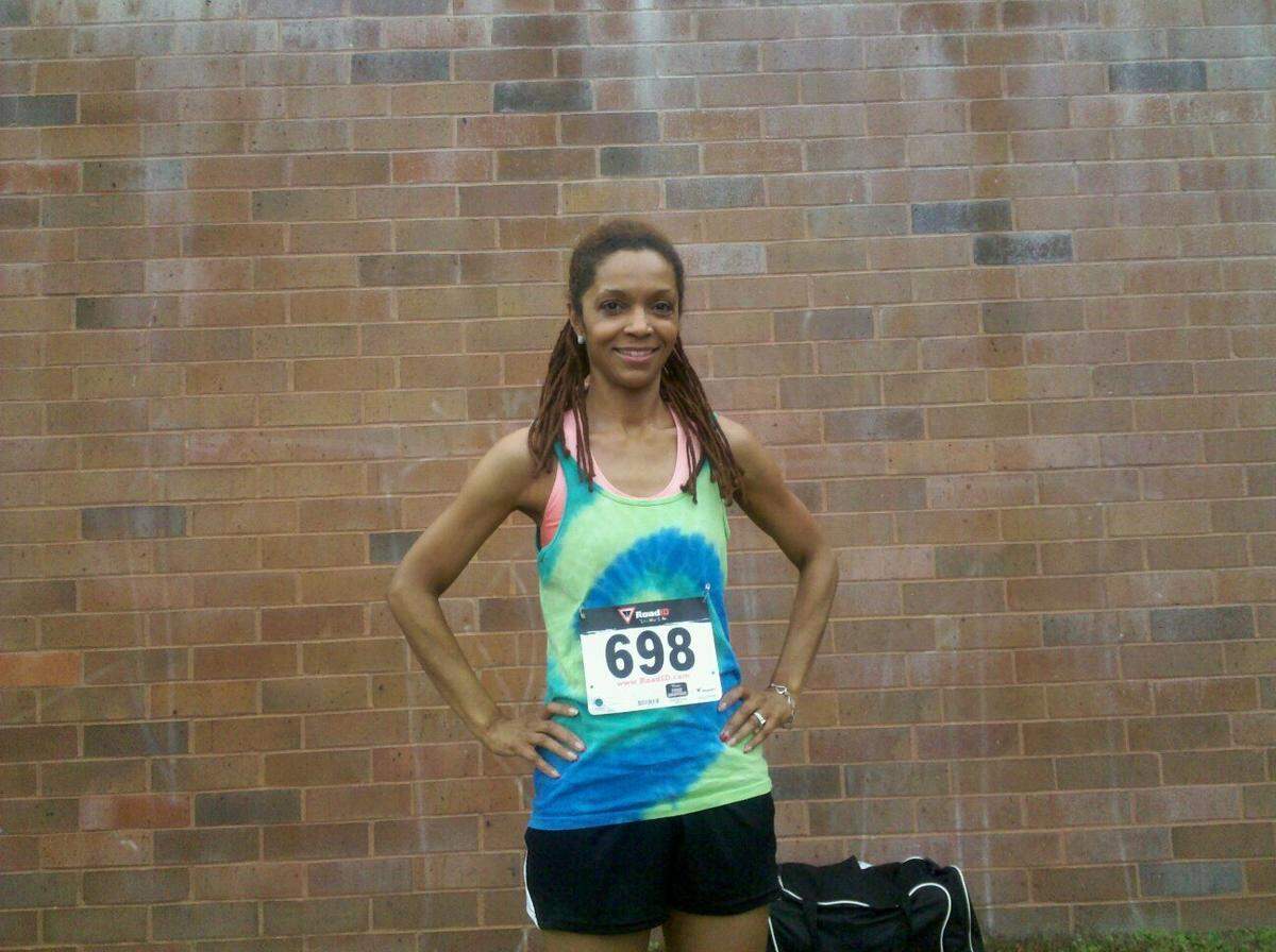 A medium, eye-level shot shows a woman with dreadlocks smiling and posing with her hands on her hips. She is wearing a green and blue tie-dye running tank top with a “698” race bib, black shorts, and a pink sports bra, standing in front of a light brown brick wall.