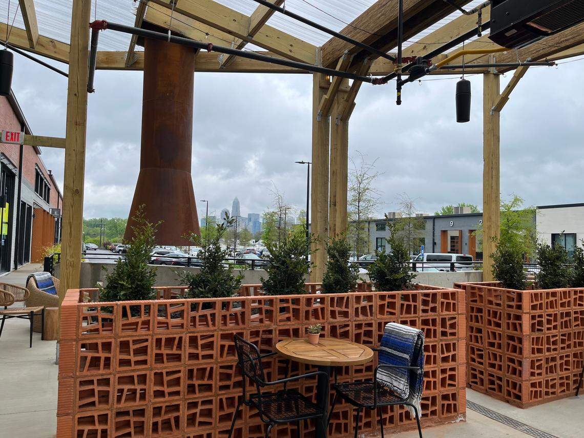 An outdoor covered patio with a rustic timber frame and a view of a city skyline in the distance under an overcast sky. In the foreground, a small round wooden table with two black metal chairs sits in front of a decorative orange terracotta breeze-block planter filled with green shrubs. A blue and white striped blanket is draped over one of the chairs, and a tall, weathered metal chimney structure stands to the left.