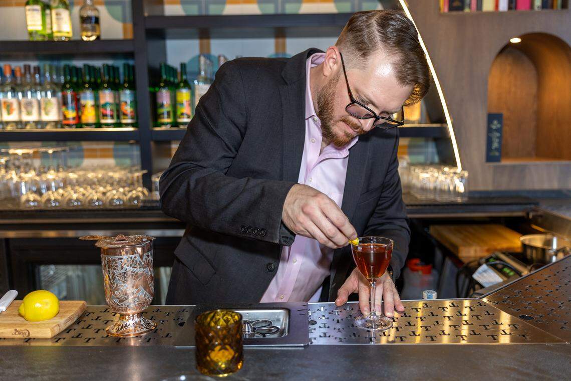 A close-up of a bartender wearing a suit jacket and pink shirt, carefully garnishing a dark amber drink in a coupe glass with a small lemon peel. A decorative copper shaker and a whole lemon are visible on the bar counter.