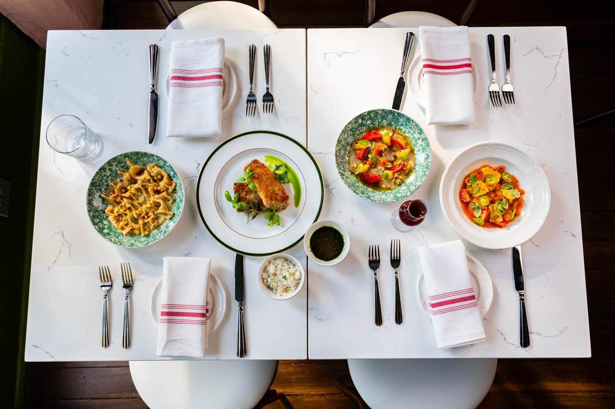 A white table is set with white plates and white napkins with red stripes, with four colorful pasta dishes in the center, two in white dishes and two in green dishes. Four white chairs are tucked under the table and silverware is at the place settings. 