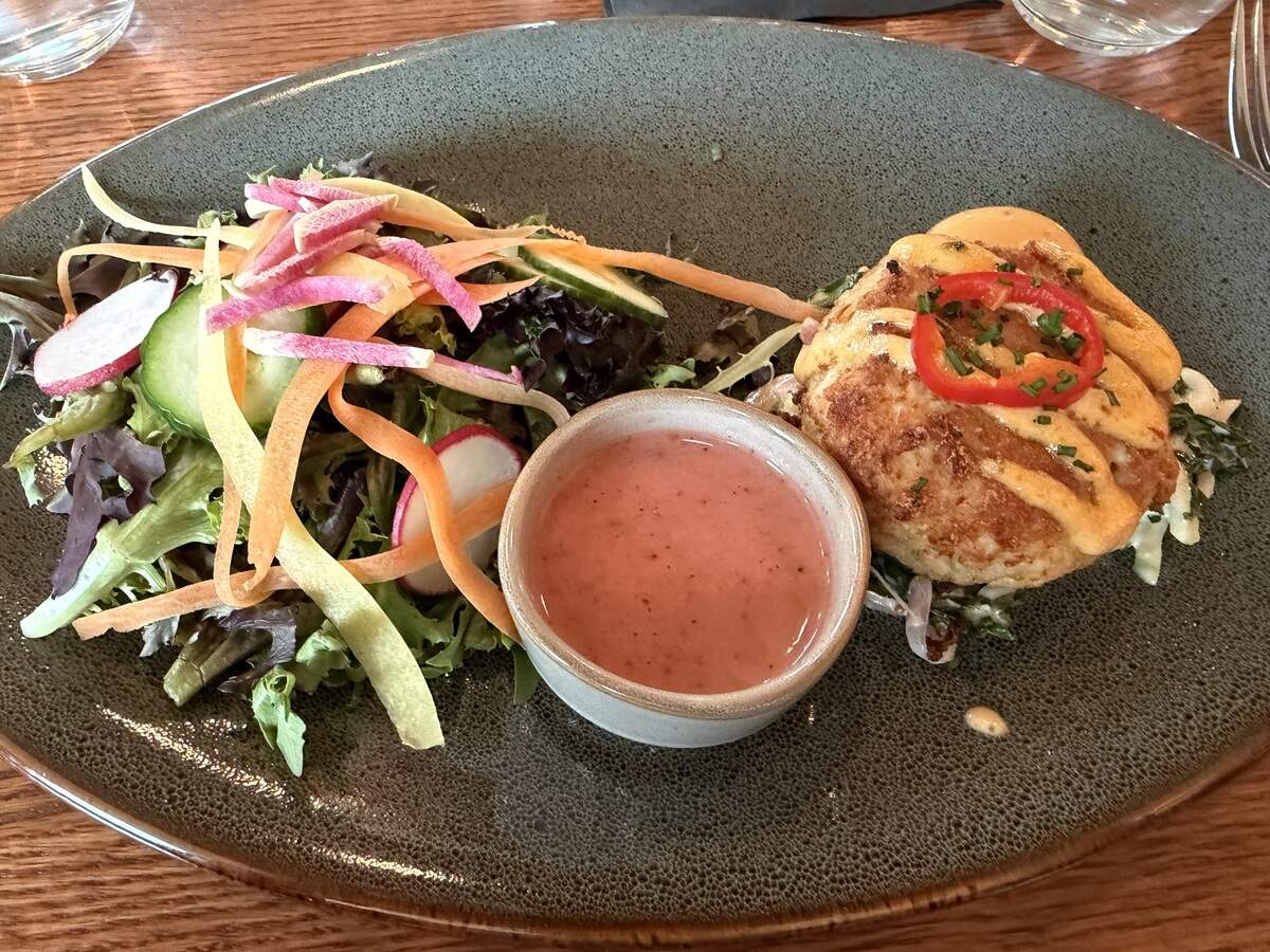 A golden-brown crab cake drizzled with sauce and garnished with a red pepper ring, served on a textured grey platter alongside a side salad with shaved carrots and radishes, and a small bowl of pink dressing.