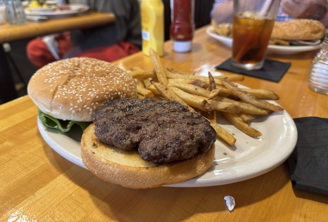 An open-faced burger served on a white plate with a thick beef patty and a side of seasoned fries. The meal is set on a wooden table in a casual dining environment.