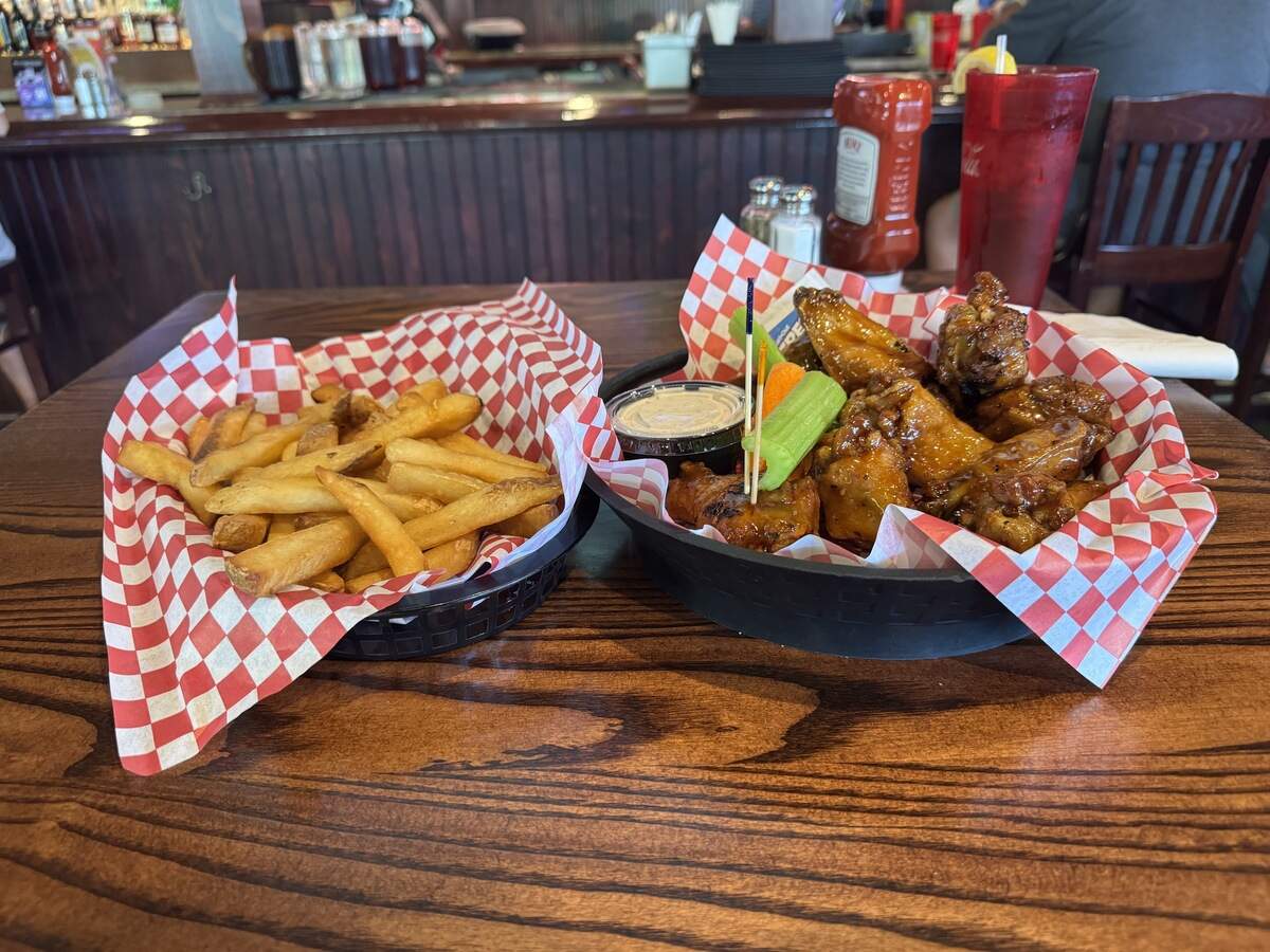 A close-up shot of pub food served in two black plastic baskets lined with red-and-white checkered paper on a dark wood table. The basket on the left is piled with golden-brown french fries. The basket on the right contains glazed barbecue chicken wings, served with celery sticks, carrot pieces, and a small plastic cup of dipping sauce. A red plastic cup with a lemon wedge and a bottle of ketchup are visible in the background.