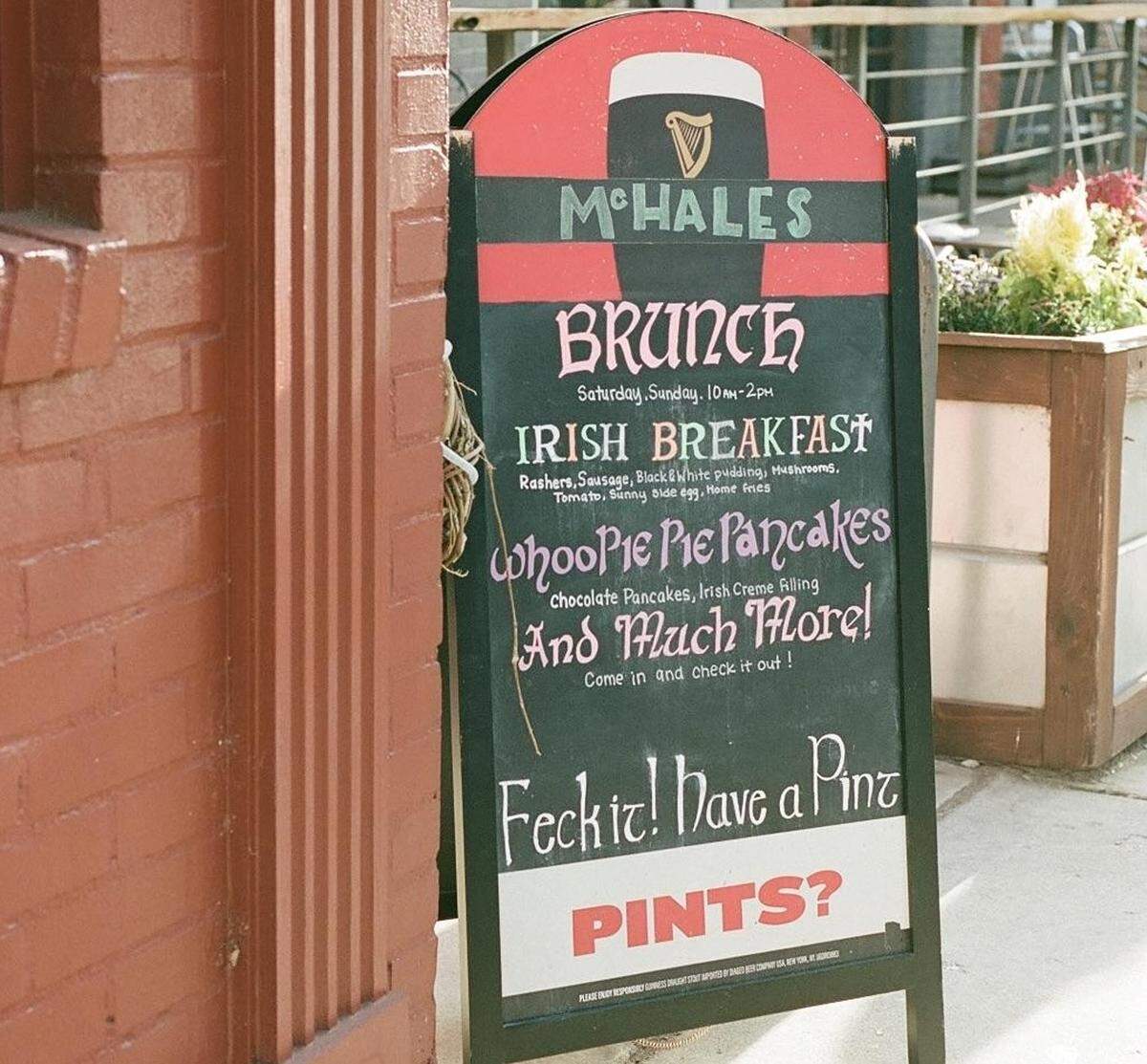 A close-up shot of a black A-frame chalkboard sign standing on a sidewalk next to a red brick wall. The sign advertises “M’HALES BRUNCH,” listing specials like “Irish Breakfast” and “Whoopie Pie Pancakes,” and concludes with the playful phrase, “Feck it! Have a Pint.”