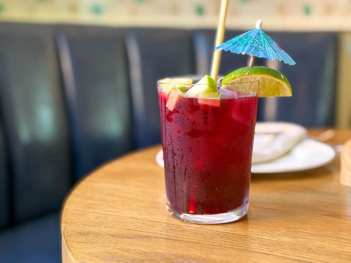 A refreshing glass of ruby-red liquid beaded with condensation, is the centerpiece of this inviting photo. Sitting on a wooden table, the drink is brimming with chopped fruit and ice, and playfully garnished with a bright lime wedge and a cheerful blue cocktail umbrella.