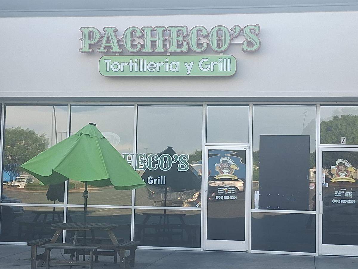 The exterior storefront of Pacheco’s Tortilleria y Grill. A sign with the restaurant’s name in green lettering hangs above large glass windows. In front, a wooden picnic table with a bright green patio umbrella sits on the sidewalk.