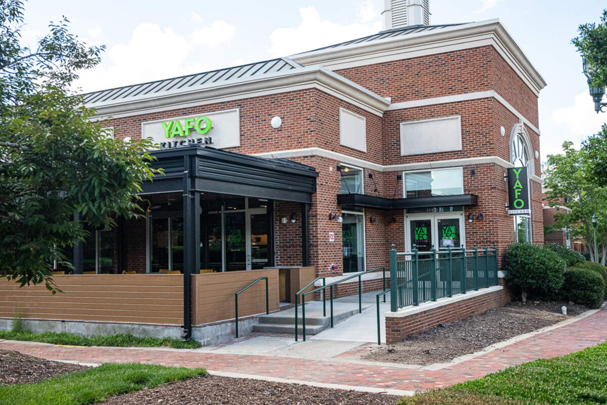 A slightly angled, wide shot of the exterior of a brick building that houses the restaurant Yafo Kitchen. The restaurant has a covered outdoor patio with a brown wood-paneled facade. A ramp and stairs with a green railing lead up to the entrance. The name “YAFO KITCHEN” is visible on several signs on the building and on a vertical sign to the right of the entrance, all in bright green letters. The building has a red brick facade with white trim, and the sidewalk is made of red brick pavers. Green trees and bushes are also visible.
