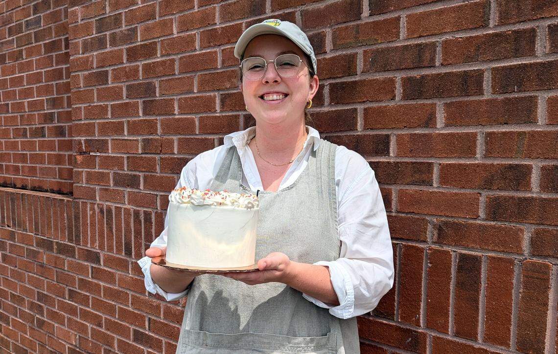 A smiling young woman, wearing a light-colored baseball cap, a white shirt, and a gray apron, stands in front of a red brick wall. She is holding a two-layer, white-frosted cake on a gold cake board. The cake is decorated with a ring of white frosting on top and is sprinkled with what appear to be tiny, colorful sprinkles. She is looking directly at the camera with a happy expression.