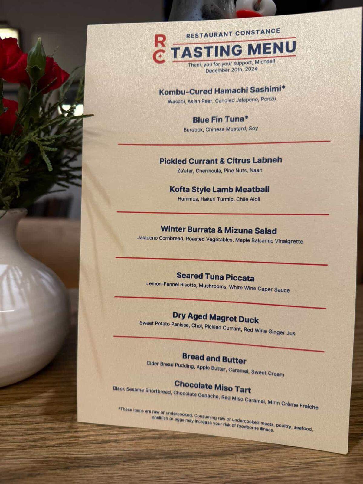 A close-up of the tasting menu from Restaurant Constance, dated December 20th, 2024, sitting on a table next to a vase of red flowers. The multi-course menu features dishes like kombu-cured hamachi, kofta style lamb meatball, seared tuna piccata and a chocolate miso tart.