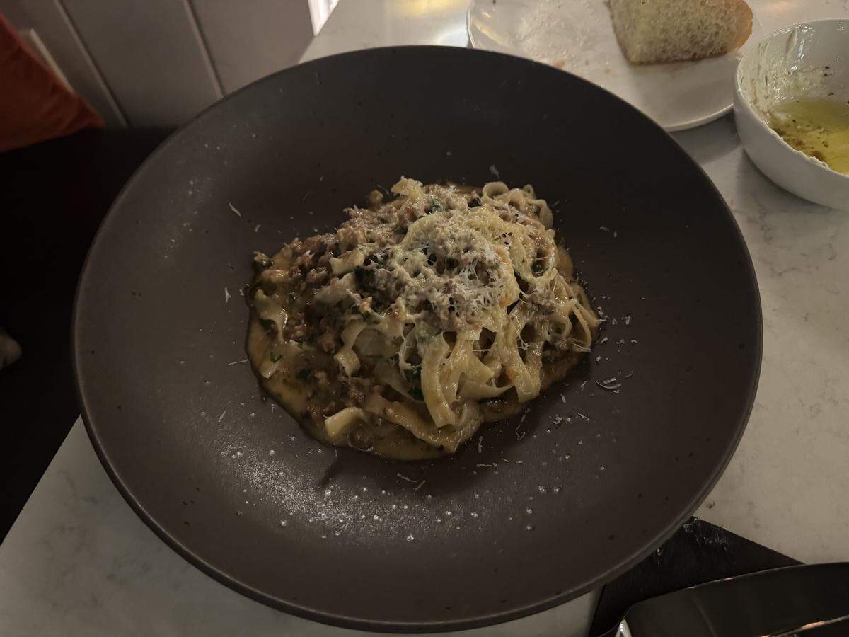 A close-up shot of a pasta dish served in a wide, matte dark-brown bowl. The dish consists of thick, ribbon-like fettuccine noodles tossed in a rich, savory meat ragu sauce, topped with a generous dusting of finely grated Parmesan cheese.