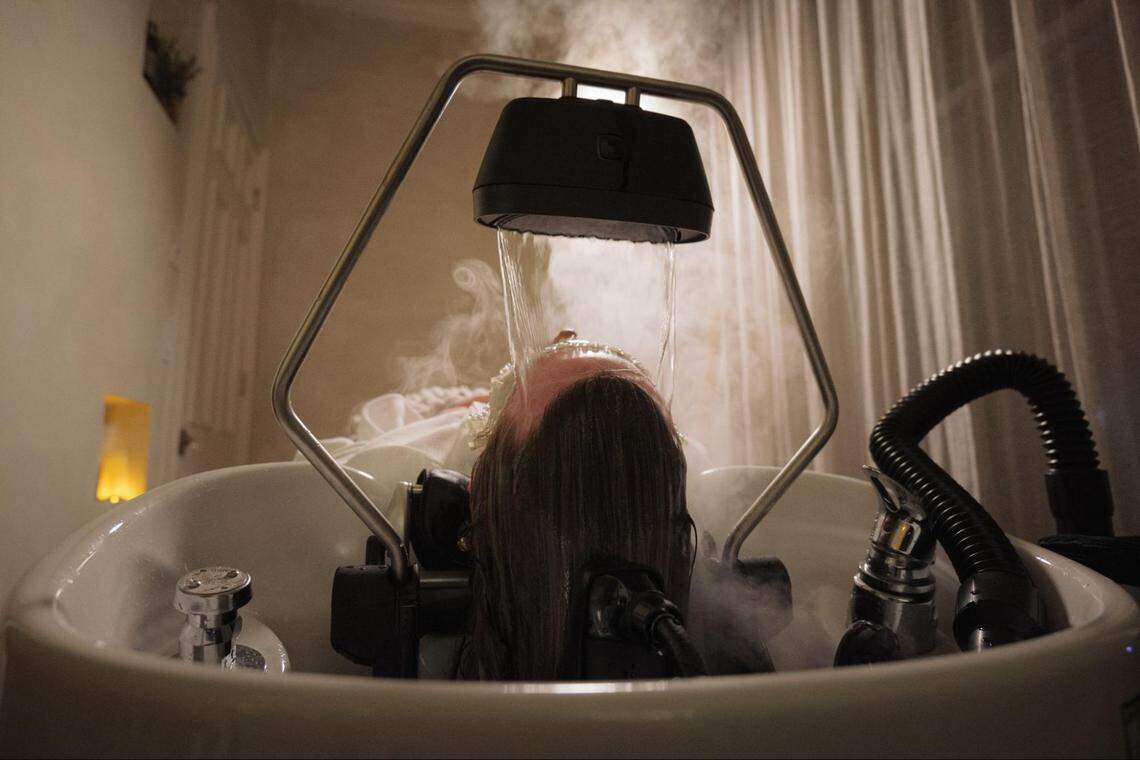 A person undergoes a “head spa” treatment where a specialized metal halo device circulates a steady, gentle stream of warm water over their forehead and scalp. Thick steam surrounds the basin and the client’s head.
