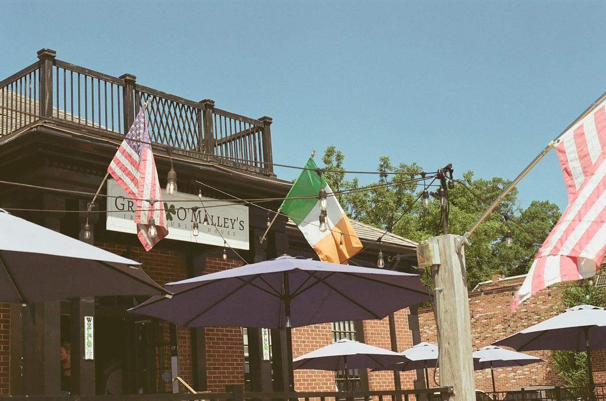 A low-angle, sunlit shot of the red brick facade of Grace O’Malley’s Public House. American and Irish flags hang above the entrance sign, and the outdoor patio in the foreground is shaded by multiple open purple umbrellas.
