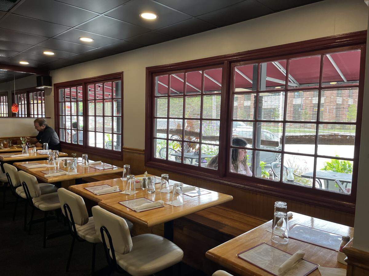 A view down a line of empty dining tables inside a restaurant. The tables are light wood and set with placemats and glasses. To the right, large multi-pane windows with dark red frames look out onto a patio area. One customer is seated at a distant table inside, and another can be seen through the window on the patio.