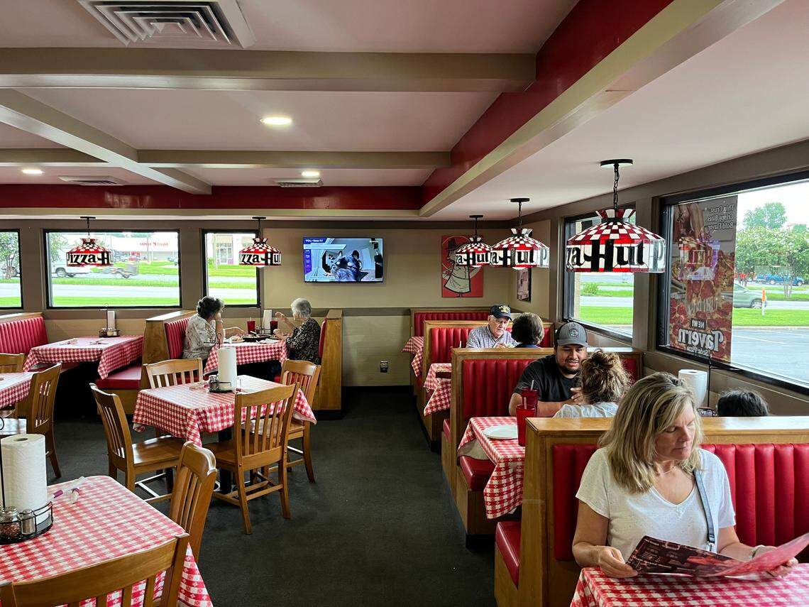 A wide shot of the interior of a retro-style Pizza Hut restaurant, featuring its classic red vinyl booths and tables with red-and-white checkered tablecloths. Iconic Tiffany-style lamps with the “Pizza Hut” logo hang over the tables. Several patrons are seated and dining in the booths, including a woman in the foreground reading a menu.