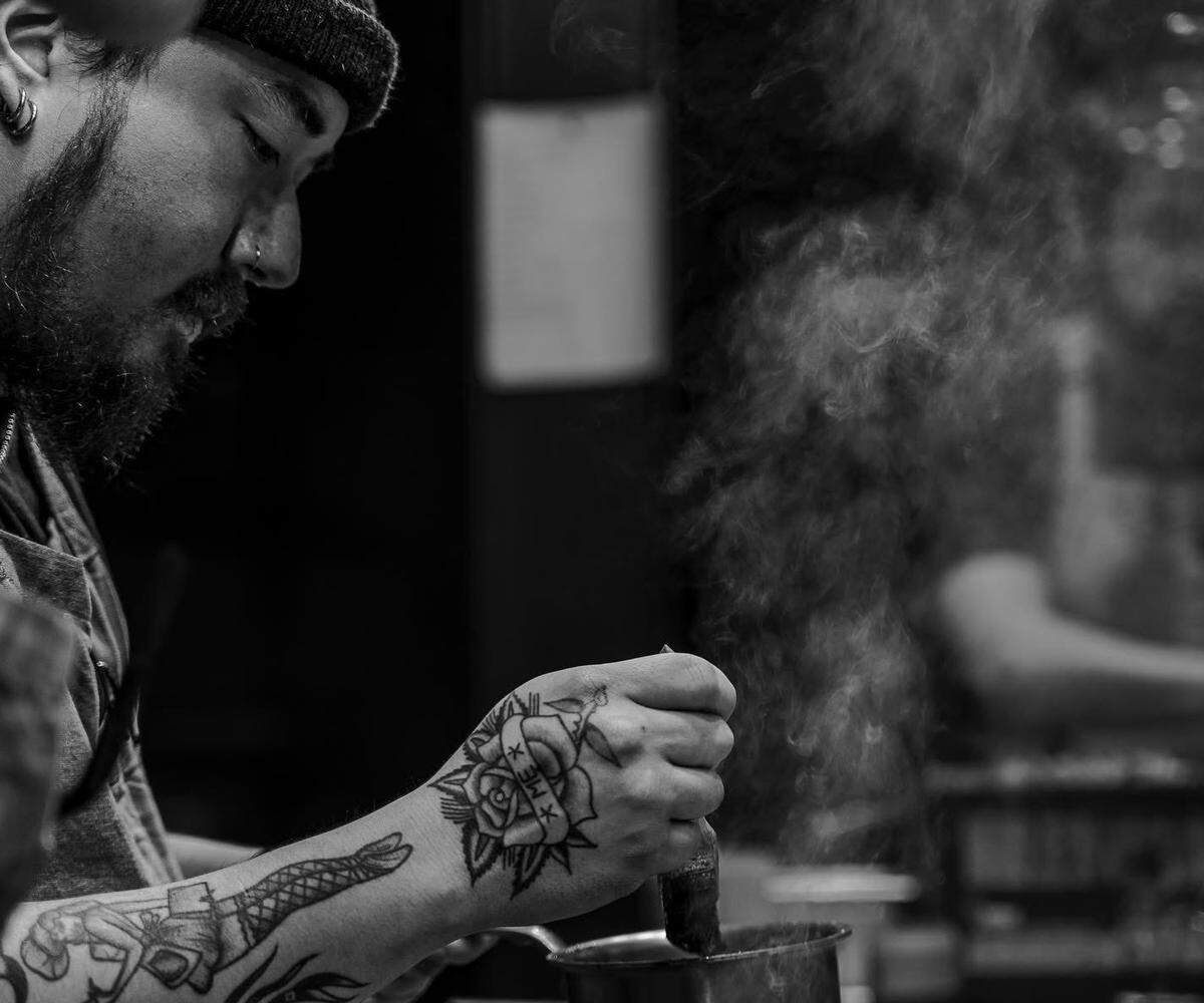 A black-and-white, profile-view photo of a chef with a beard and a beanie hat focusing on a pot. The chef has several tattoos, including a prominent rose on the back of the hand and a figure on the forearm. Thick steam or smoke rises from the pot the chef is stirring in a dark, atmospheric setting.
