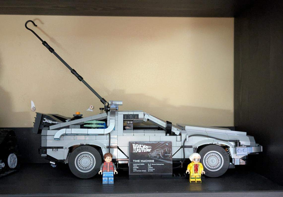 A side-profile shot of the LEGO “Back to the Future” Time Machine model, displayed on a dark shelf. The gray DeLorean build includes the long lightning rod hook attached to the back, arcing up and forward. In front of the car stand the LEGO minifigures of Marty McFly and Doc Brown, flanking a black display plaque with the movie’s logo and car details.