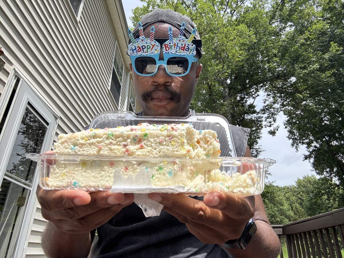 A close-up shot of a person wearing novelty “Happy Birthday” sunglasses with candle decorations, holding up a clear plastic container containing a large slice of confetti cake with white frosting and rainbow sprinkles. The background shows the side of a house with light-colored siding and green trees.