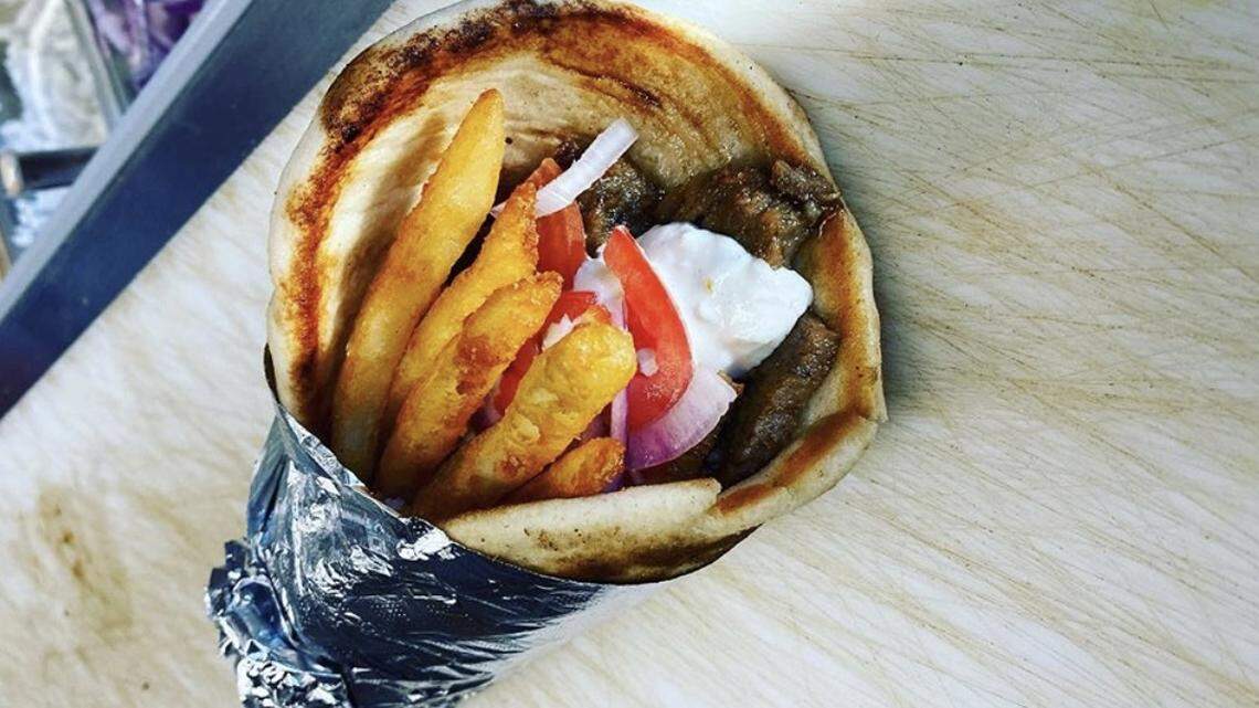 A close-up, top-down photograph of a gyro. The soft pita bread, wrapped in foil at its base, is generously filled with sliced gyro meat, french fries, tomato, and onion, and is topped with a large dollop of white tzatziki sauce.