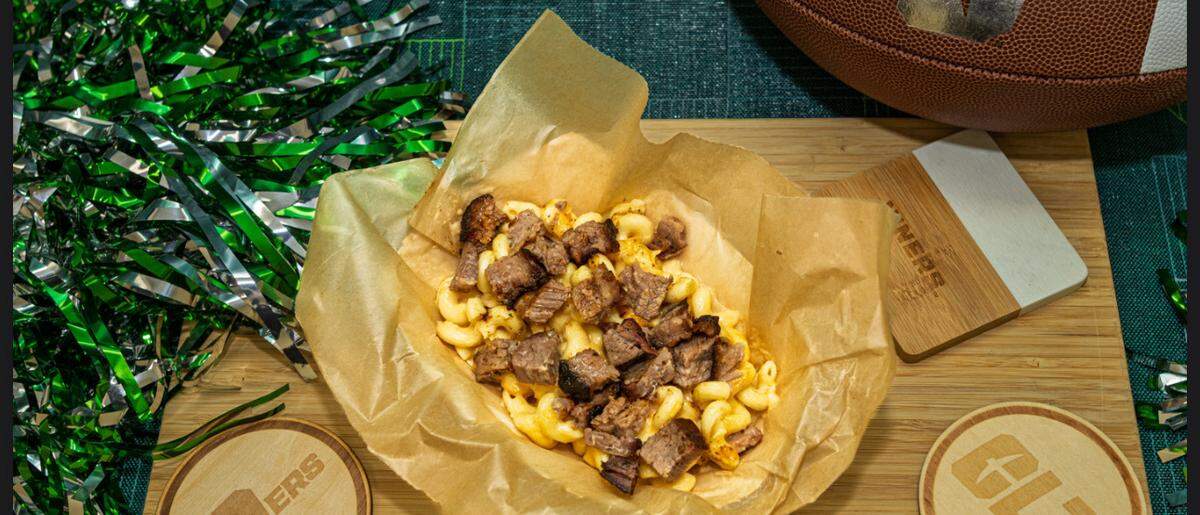 A dish of macaroni and cheese topped with pieces of brisket. The food is served in a brown paper bowl on a wooden cutting board. Surrounding the food are football-themed items, including a football, a green and silver pom-pom, and two wooden coasters with “9ERS” and “CLT” logos.
