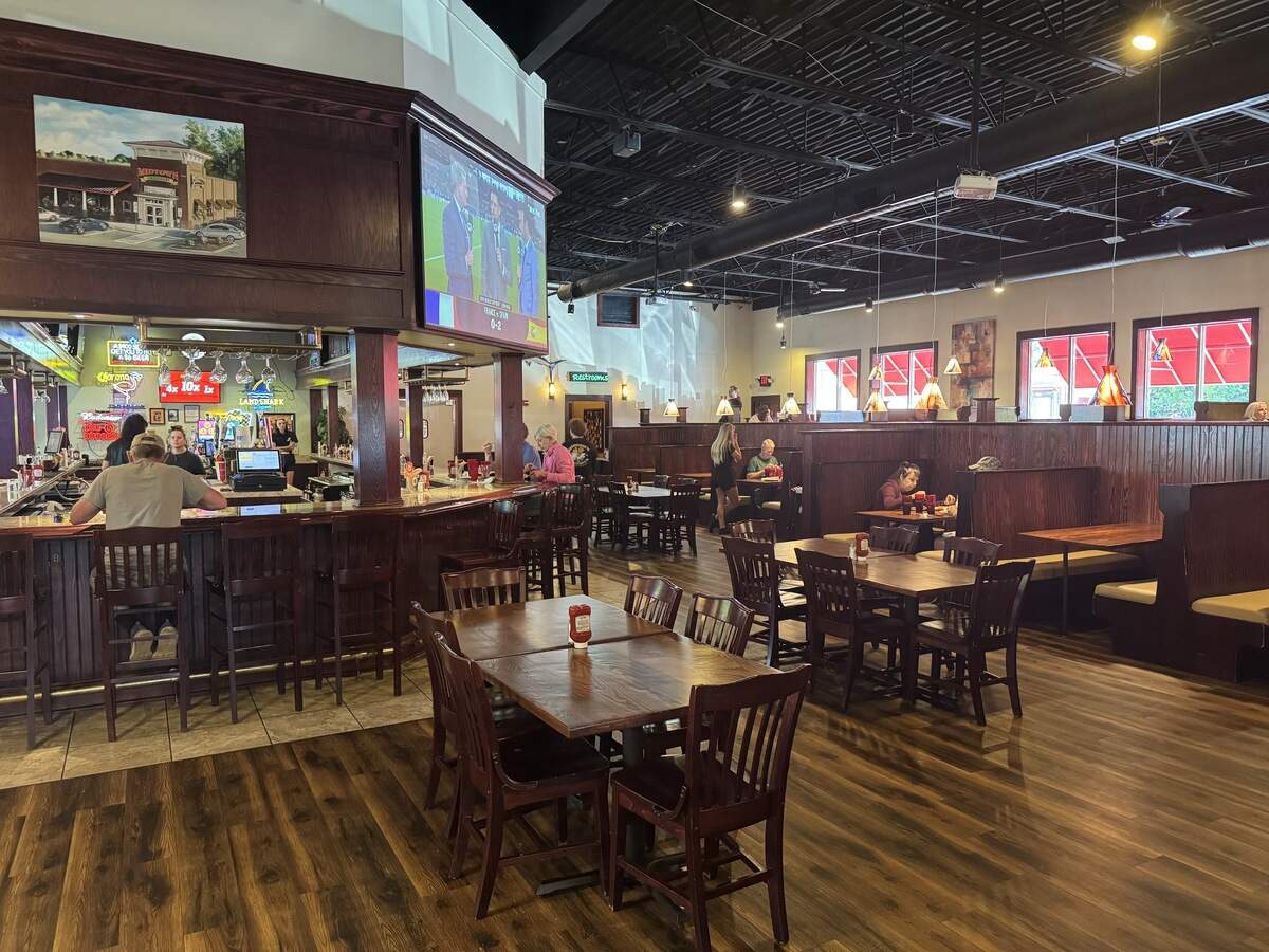 The spacious interior of a warm, casual sports bar and grill. The room features dark wood-plank flooring, a high exposed industrial ceiling painted black, and numerous wooden tables and chairs. On the left, a large u-shaped dark wood bar is surrounded by barstools, with a flat-screen TV overhead showing a sports broadcast. High-walled wooden booths line the right wall under windows with red awnings.
