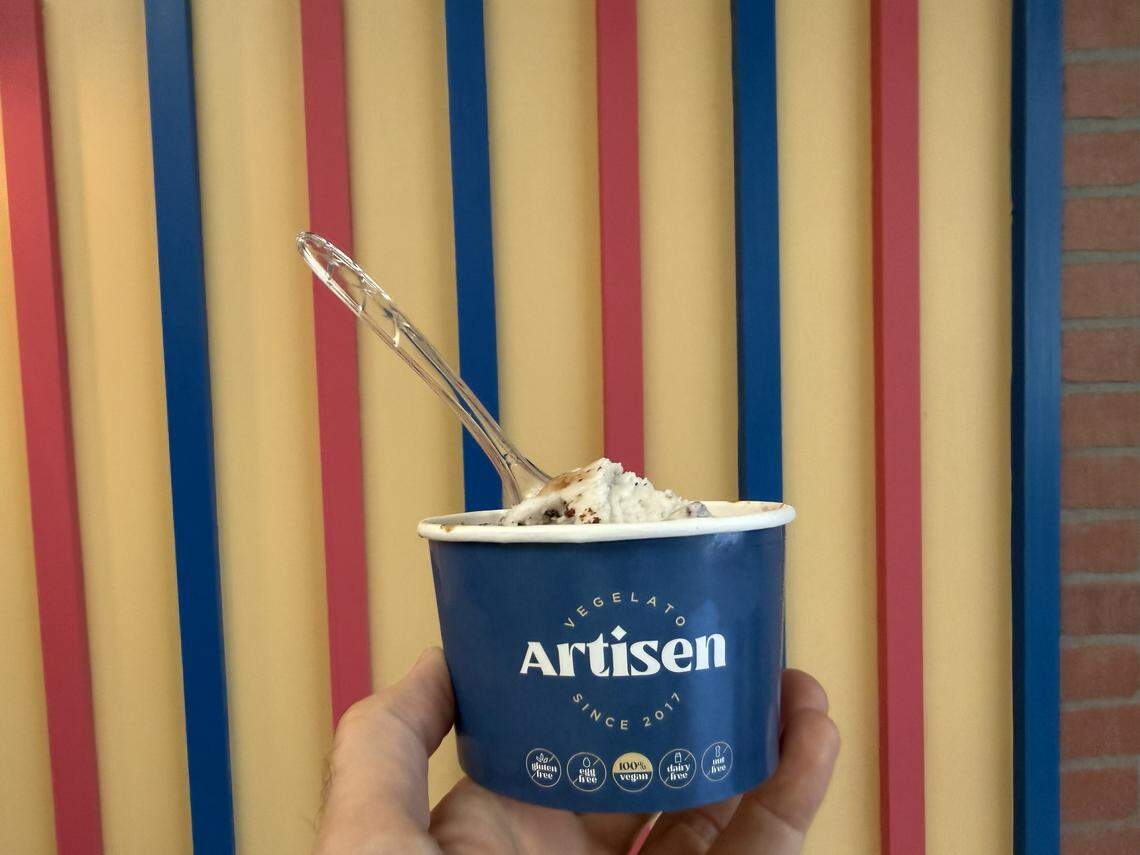 A hand holds a blue paper cup filled with a scoop of white vegelato and a clear plastic spoon. The cup features the Artisen logo and “Since 2017” in white lettering, with small icons at the bottom highlighting its allergen-friendly qualities, including “100% Vegan.”
