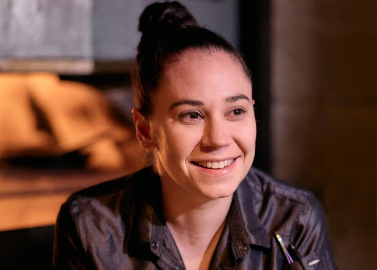 A close-up portrait of a smiling chef with hair pulled back into a high bun, is wearing a dark, iridescent grey button-up shirt or chef’s coat. The background is softly blurred with warm, ambient lighting, suggesting a restaurant or kitchen setting.