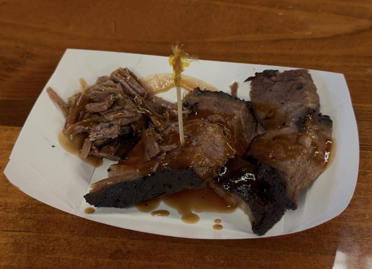 A close-up shot of a small paper food boat sitting on a polished wooden surface. Inside the boat are multiple thick slices of tender, slow-cooked beef brisket with a dark, caramelized bark on the edges. The meat is generously drizzled with a glossy brown barbecue sauce, and a single wooden toothpick with a decorative frill is inserted into the center slice.