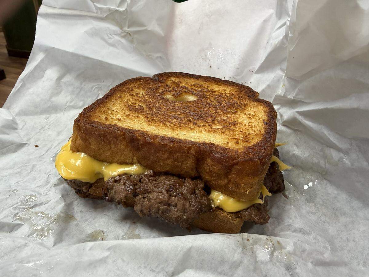 A close-up, slightly high-angle shot of a whole patty melt sandwich sitting on a piece of crumpled white deli paper. The sandwich consists of a thick, seasoned beef patty topped with gooey, melted yellow American cheese, all served between two thick slices of perfectly golden-brown, butter-toasted Texas toast.