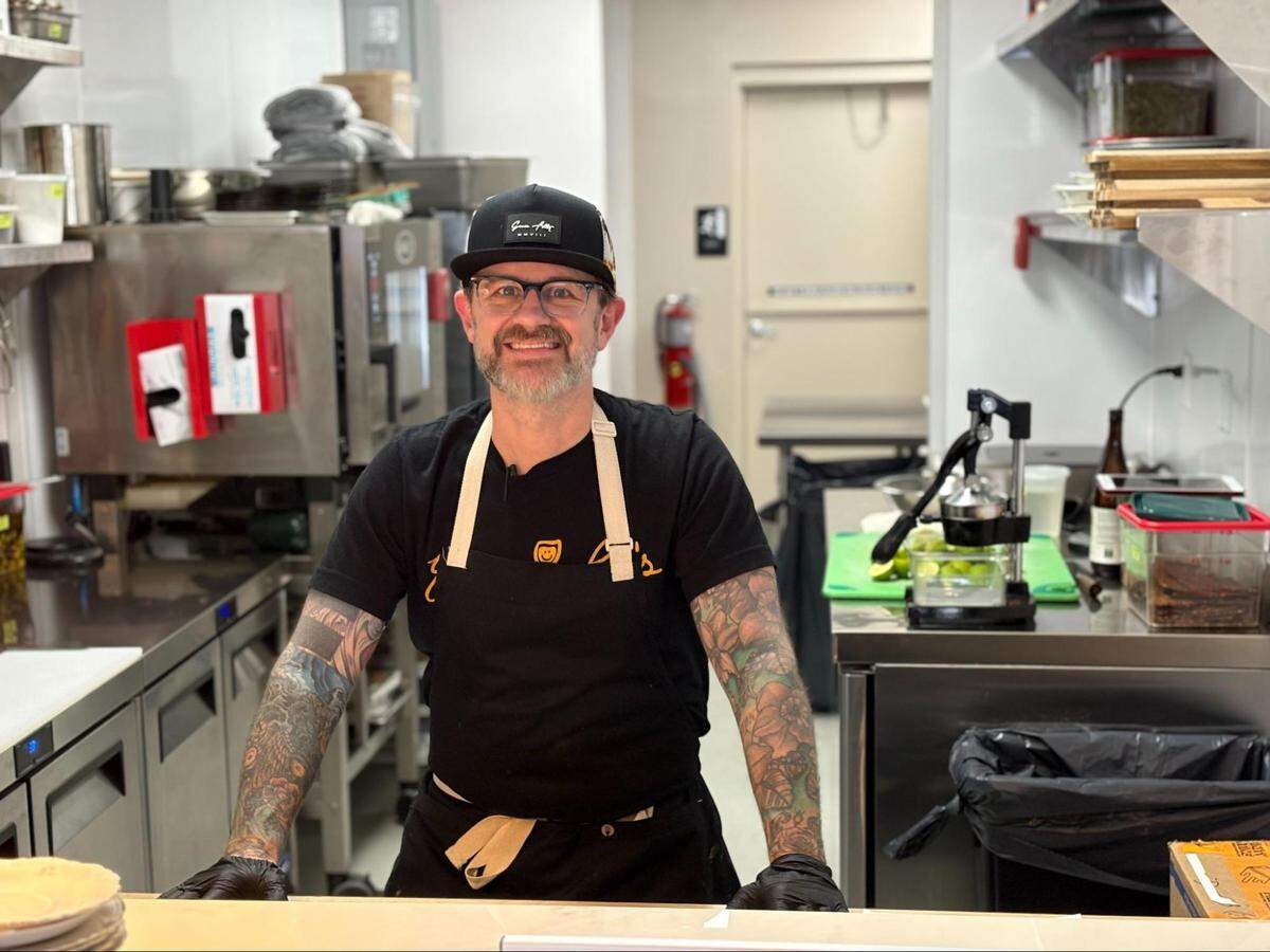 A medium shot of a smiling chef with glasses, a salt-and-pepper beard, and heavily tattooed arms standing in a commercial kitchen. The chef is wearing a black t-shirt, a black apron with tan straps, and a black baseball cap with a white logo. The background shows professional kitchen equipment, including stainless steel prep tables, a citrus juicer, and shelving.
