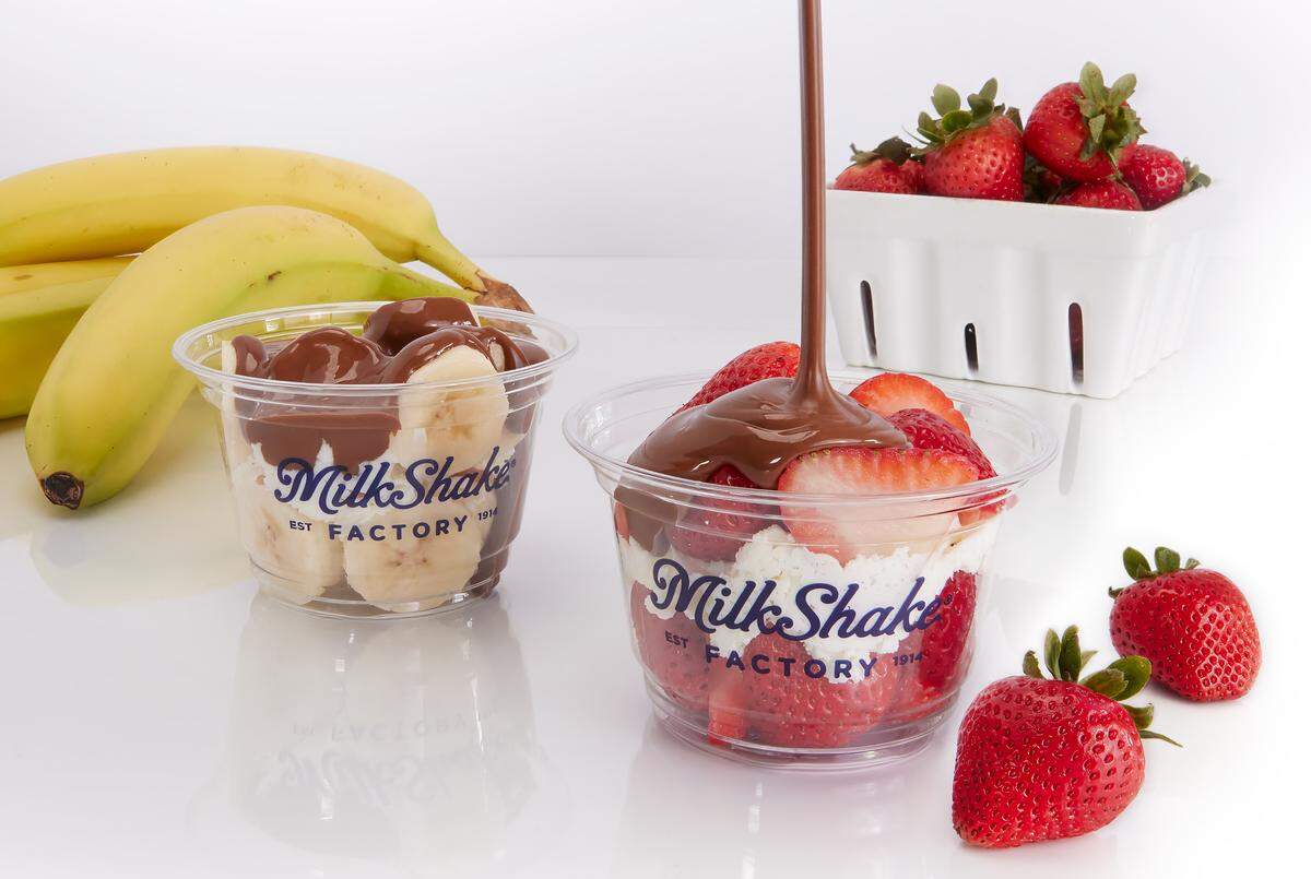 A close-up shot shows two Milkshake Factory molten chocolate cups with fresh fruit. One cup contains bananas and is topped with chocolate, while the other contains strawberries as a stream of chocolate is being poured onto it.