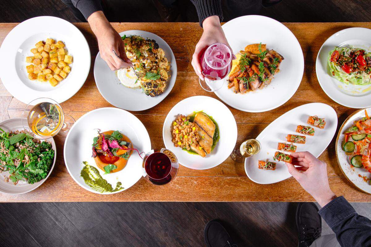 A high-angle, top-down photograph of a long wooden dining table filled with an elaborate spread of gourmet dishes and drinks at Guard and Grace. Two people are partially visible, their hands reaching into the frame to interact with the meal.