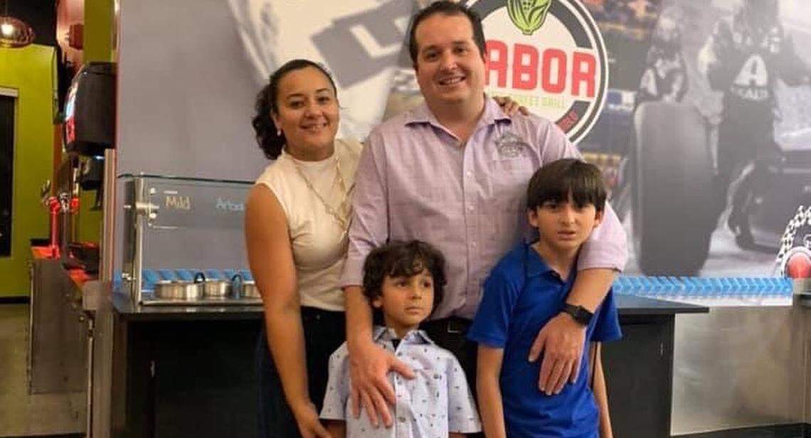 A warmly lit family portrait of a man, a woman and two young children. They are standing close together in a fast-casual restaurant setting, with a beverage and sauce station to their left and a branded mural on the wall behind them.