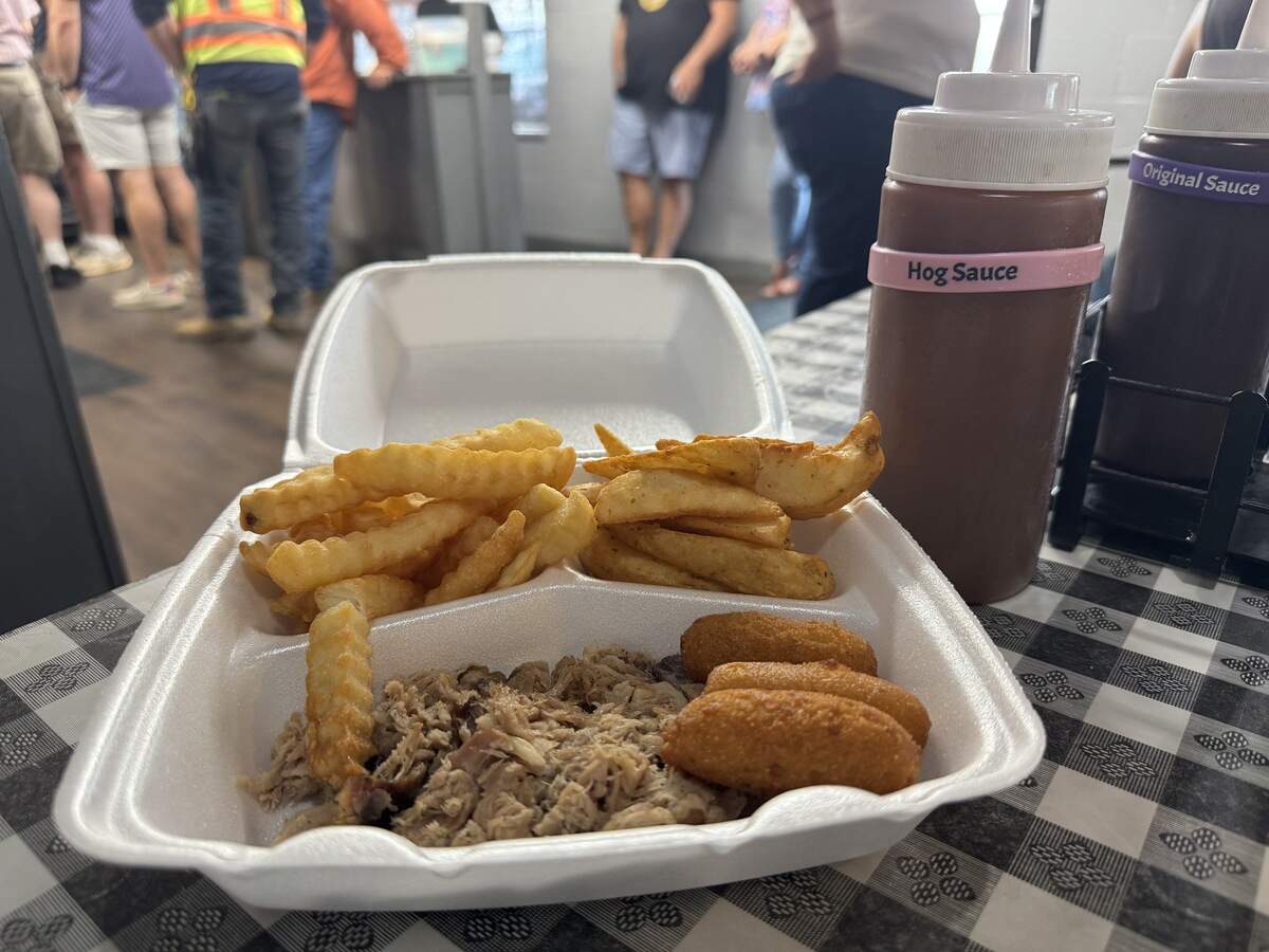 A close-up shot of a barbecue meal served in a white styrofoam container on a black-and-white checkered tablecloth. The platter includes a serving of shredded pulled pork, three golden-brown hushpuppies, crinkle-cut french fries, and potato wedges. In the background, a squeeze bottle labeled “Hog Sauce” sits next to a bottle labeled “Original Sauce,” with a blurred view of customers standing in line.