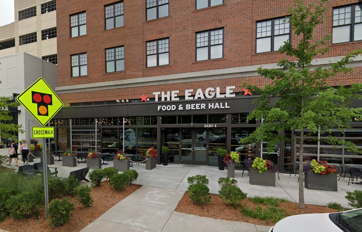 A street-level photo of a red brick building with large windows, housing a restaurant called The Eagle Food & Beer Hall. The restaurant has an outdoor patio with tables and planters filled with flowers.