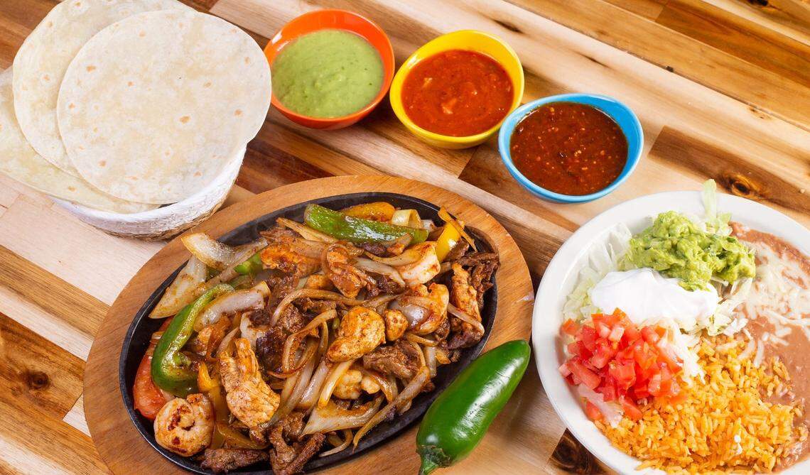 A top-down shot of a sizzling platter of chicken and steak fajitas on a wooden surface. The skillet is filled with grilled meat, onions, and green bell peppers. A roasted jalapeño sits on the side. The meal is accompanied by a white plate with Spanish rice, refried beans, guacamole, sour cream, and pico de gallo. A basket of flour tortillas and three colorful bowls of different salsas (green, red, and chunky) are also on the table.