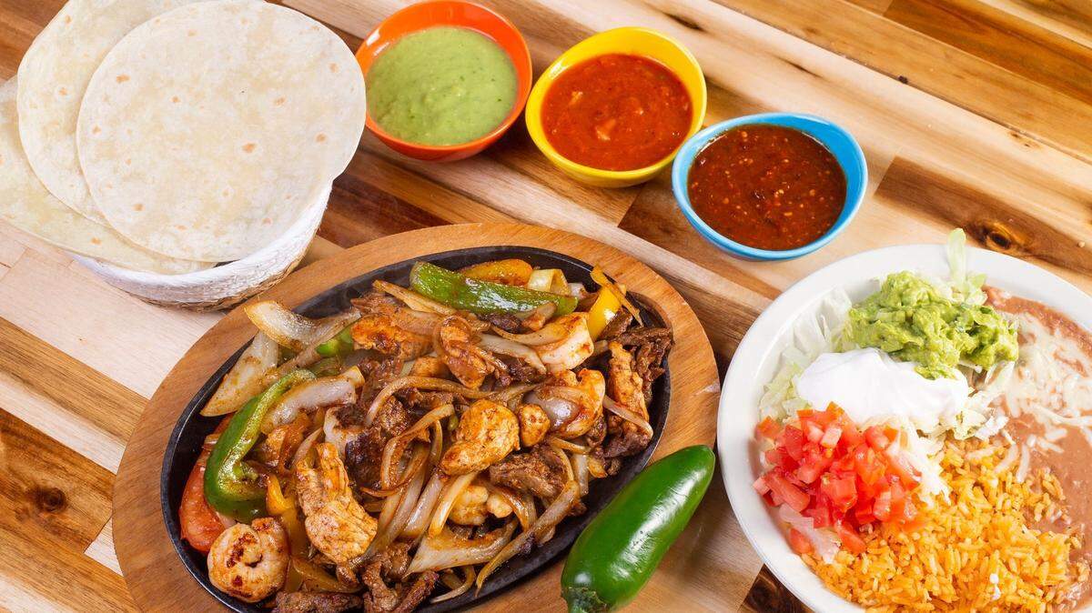A top-down shot of a sizzling platter of chicken and steak fajitas on a wooden surface. The skillet is filled with grilled meat, onions, and green bell peppers. A roasted jalapeño sits on the side. The meal is accompanied by a white plate with Spanish rice, refried beans, guacamole, sour cream, and pico de gallo. A basket of flour tortillas and three colorful bowls of different salsas (green, red, and chunky) are also on the table.