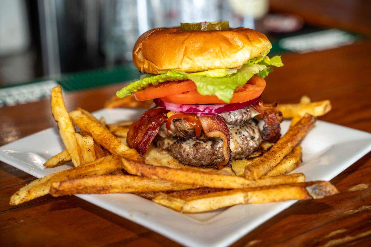 A double bacon cheeseburger topped with lettuce, tomato, red onion, and a pickle, served with a side of seasoned fries on a white plate.