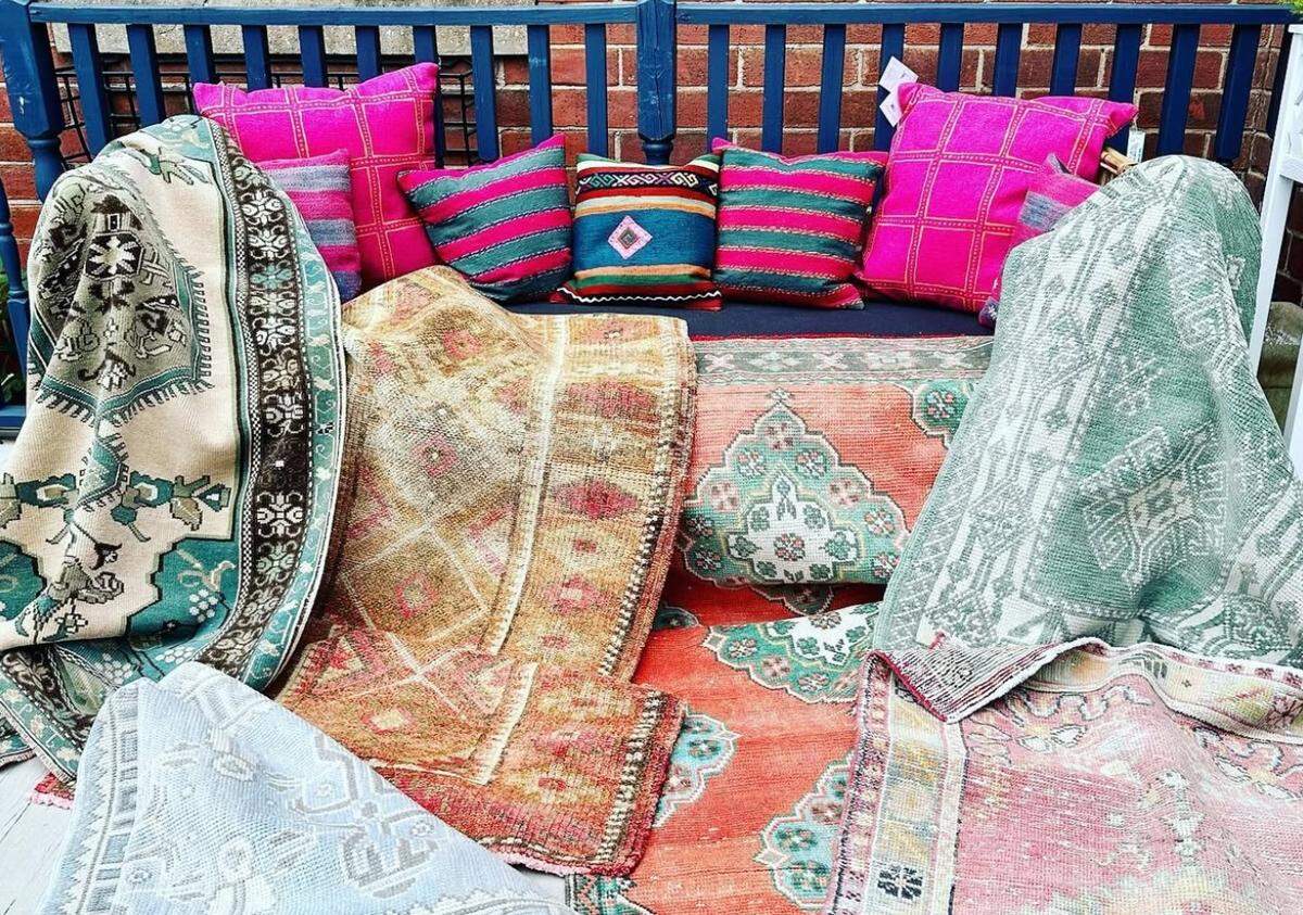 An abundant outdoor display of rugs and pillows against a blue railing. The foreground is a cascade of overlapping, vintage-style rugs with intricate, faded geometric patterns in reds, oranges, and greens. Behind the rugs, a line of brightly colored pillows, mostly hot pink and striped, are propped up for display.