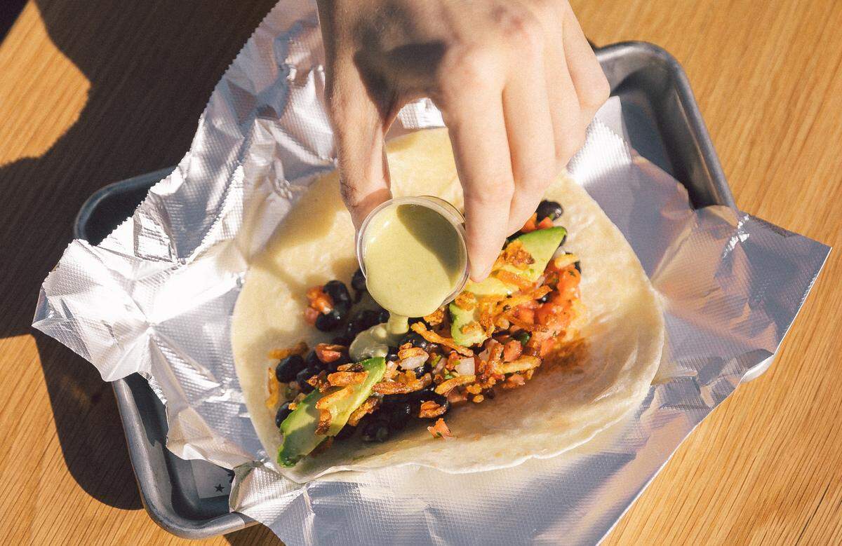 A hand pours a creamy green salsa from a plastic ramekin onto a taco loaded with black beans, avocado slices, and pico de gallo. The taco is served on crinkled silver foil over a small metal tray on a light wood table.