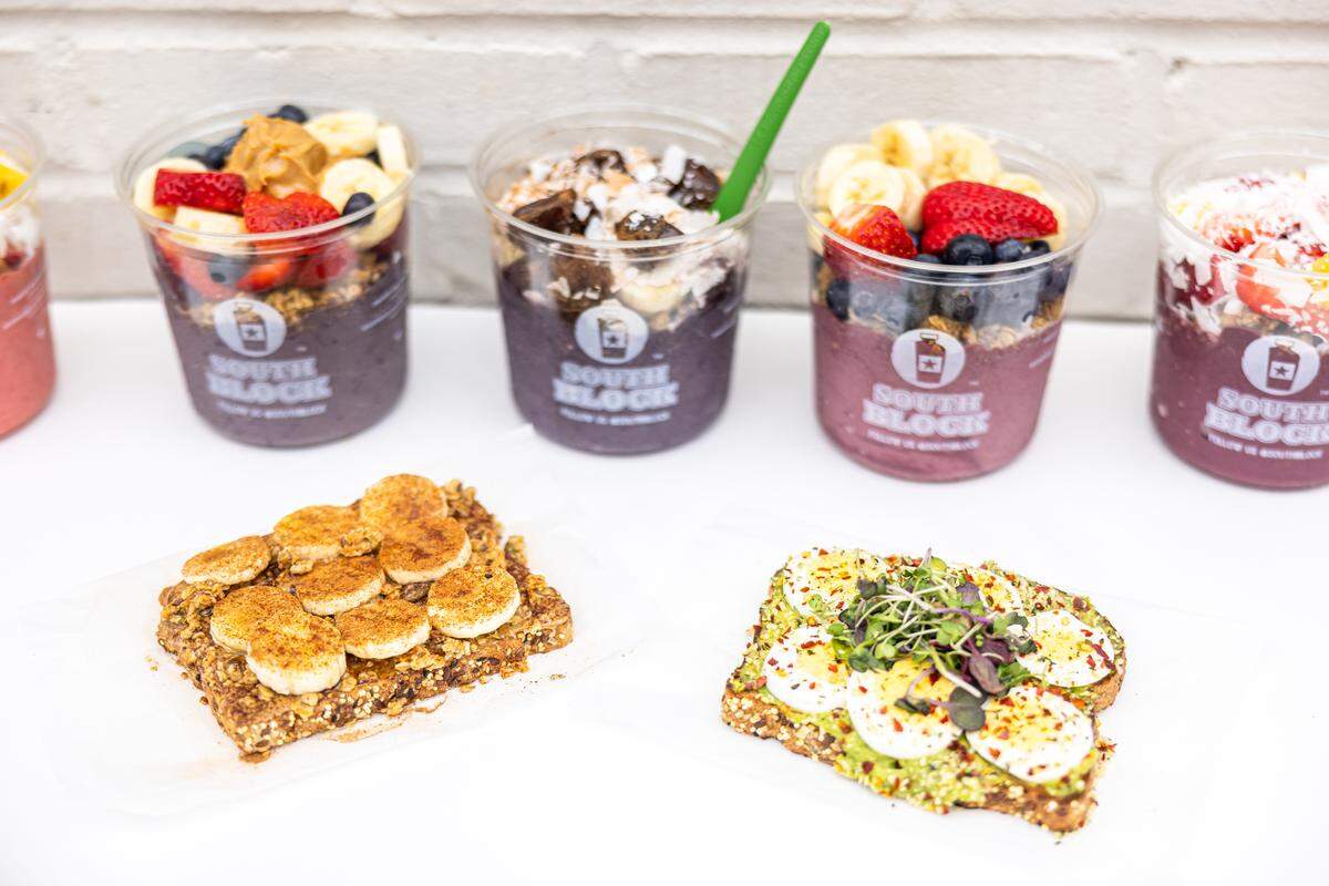 A bright, high-angle shot featuring four South Block acai bowls in clear plastic cups and two slices of loaded toast on a white surface. The acai bowls are topped with fresh fruits like bananas, strawberries, and blueberries, as well as peanut butter and coconut flakes. One slice of toast is topped with sliced bananas and cinnamon, while the other features smashed avocado, sliced hard-boiled eggs, microgreens, and red pepper flakes.