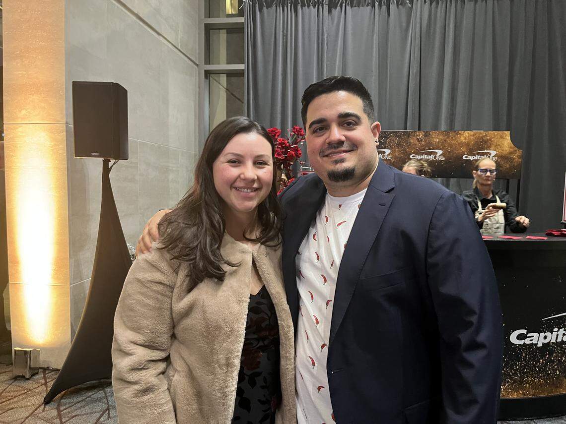 A medium, eye-level shot of a couple smiling and posing for a photo at an indoor event. The person on the left is wearing a light-tan fuzzy jacket. The one on the right has an arm around the other and is wearing a dark blue blazer over a white t-shirt with a red chili pepper print. In the background, a “Capital One” branded bar is visible.