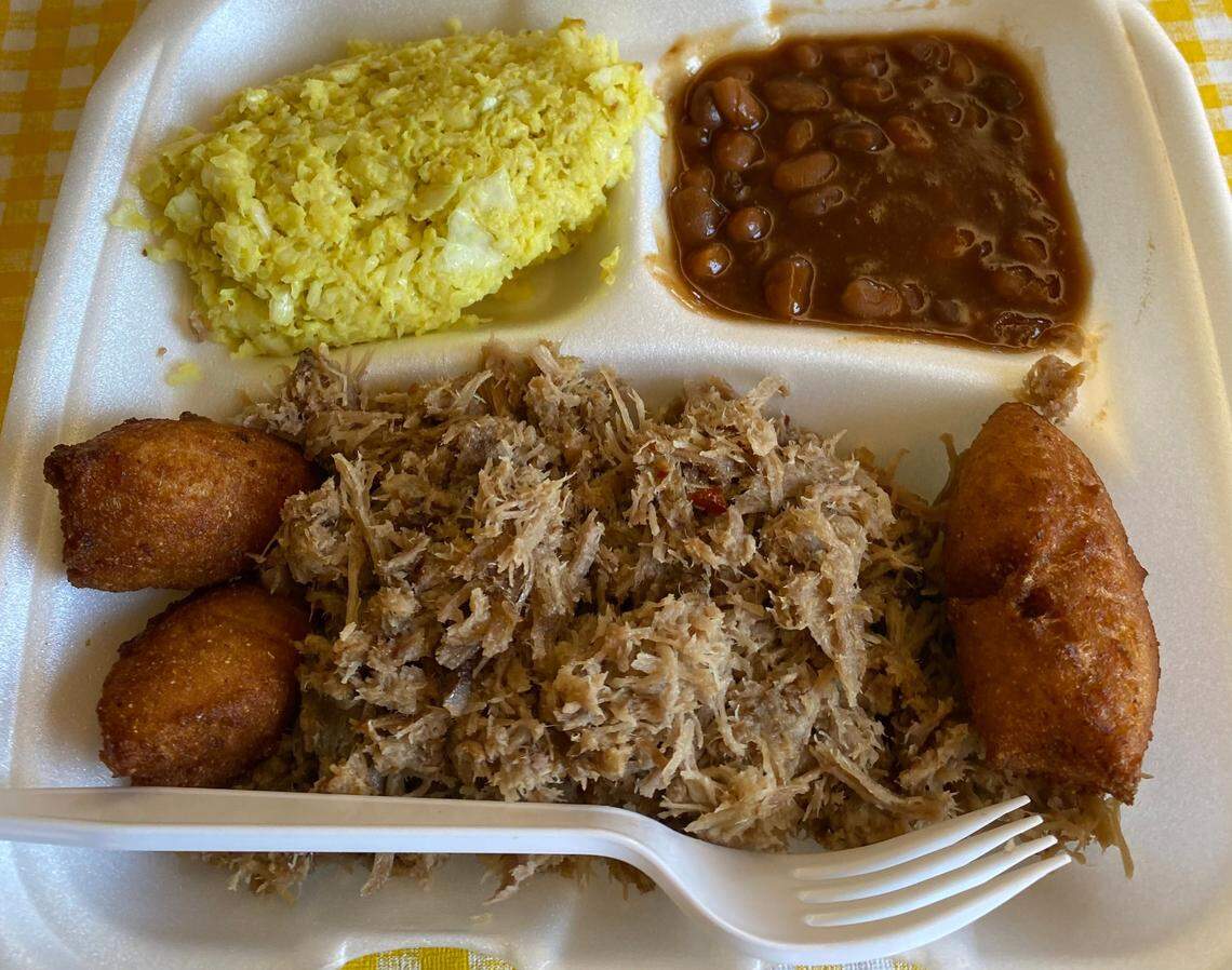 An overhead shot captures a three-compartment styrofoam container filled with a barbecue meal on a checkered tablecloth. The largest section contains a mound of chopped pork barbecue. Smaller sections hold baked beans and a yellow mustard-based side salad. Four crispy hush puppies accompany the meal.