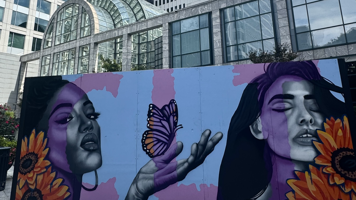 A wide shot of a beautiful street art mural on a temporary wall in a city plaza. The painting shows two large portraits of women with sunflowers near their shoulders. Between them, a hand holds a Monarch butterfly. The artist’s signature, Nony, is visible. Behind the mural stands a large, modern building with a glass atrium.