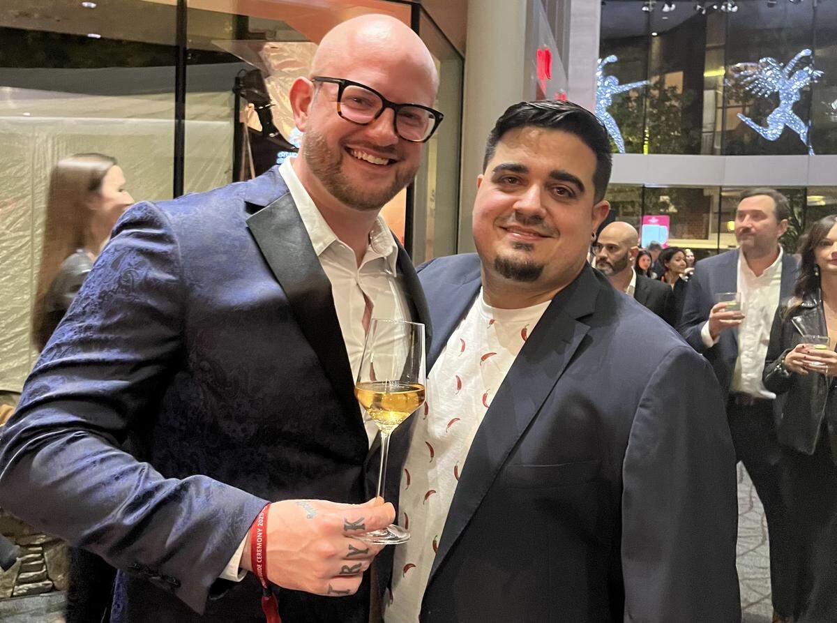 A close-up shot of two chefs smiling and posing together at an indoor event. The one on the left, who is bald and wears glasses, is in a blue patterned blazer and holds a glass of white wine. The one on the right, wearing a dark blue blazer over a t-shirt with a red chili pepper print, has an arm around the other person’s shoulder. The background is a crowded reception area with large windows.