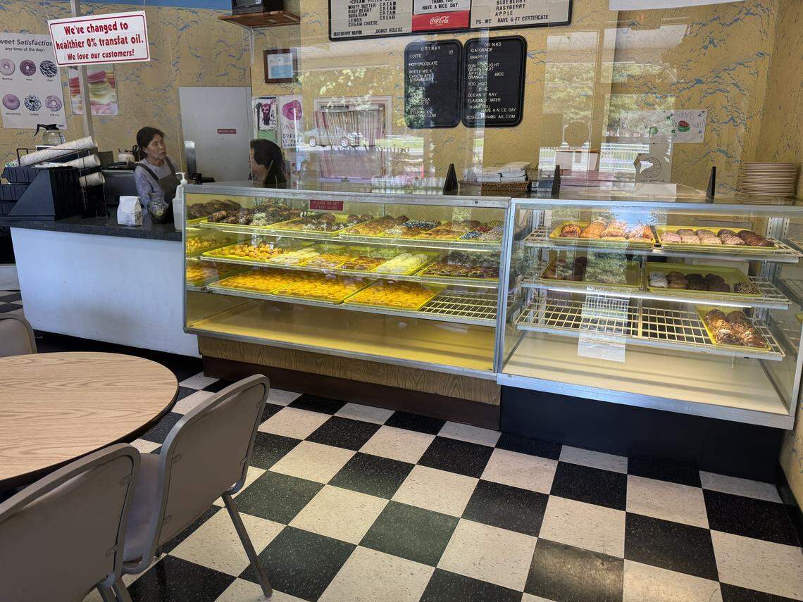 The interior of a welcoming, old-fashioned donut shop. A large glass bakery case displays rows of colorful donuts. The shop has a distinctive black and white checkered floor and yellow walls. Behind the counter, two employees are working.