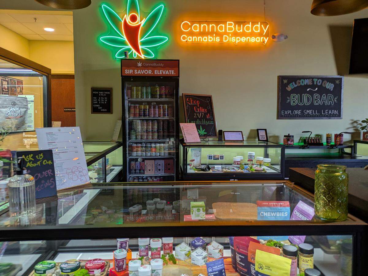 An interior view of CannaBuddy Cannabis Dispensary. A bright neon sign featuring a stylized orange figure inside a green leaf hangs above a “Bud Bar” chalkboard. Glass display cases in the foreground are filled with various cannabis products, edibles, and flower specials, while a refrigerated cooler holds infused beverages.